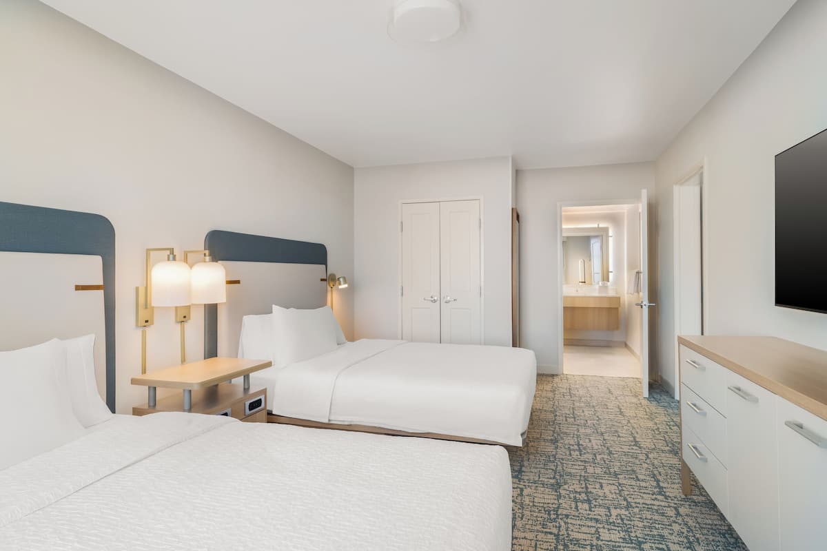 A hotel room with two beds, a desk, a TV, and a bathroom visible through an open door.