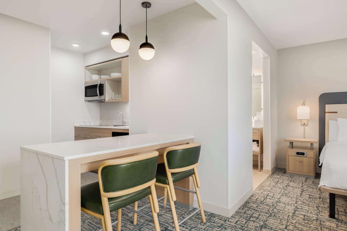 A hotel room with a kitchen area, a dining table with two chairs, a bed, and a nightstand with a lamp.