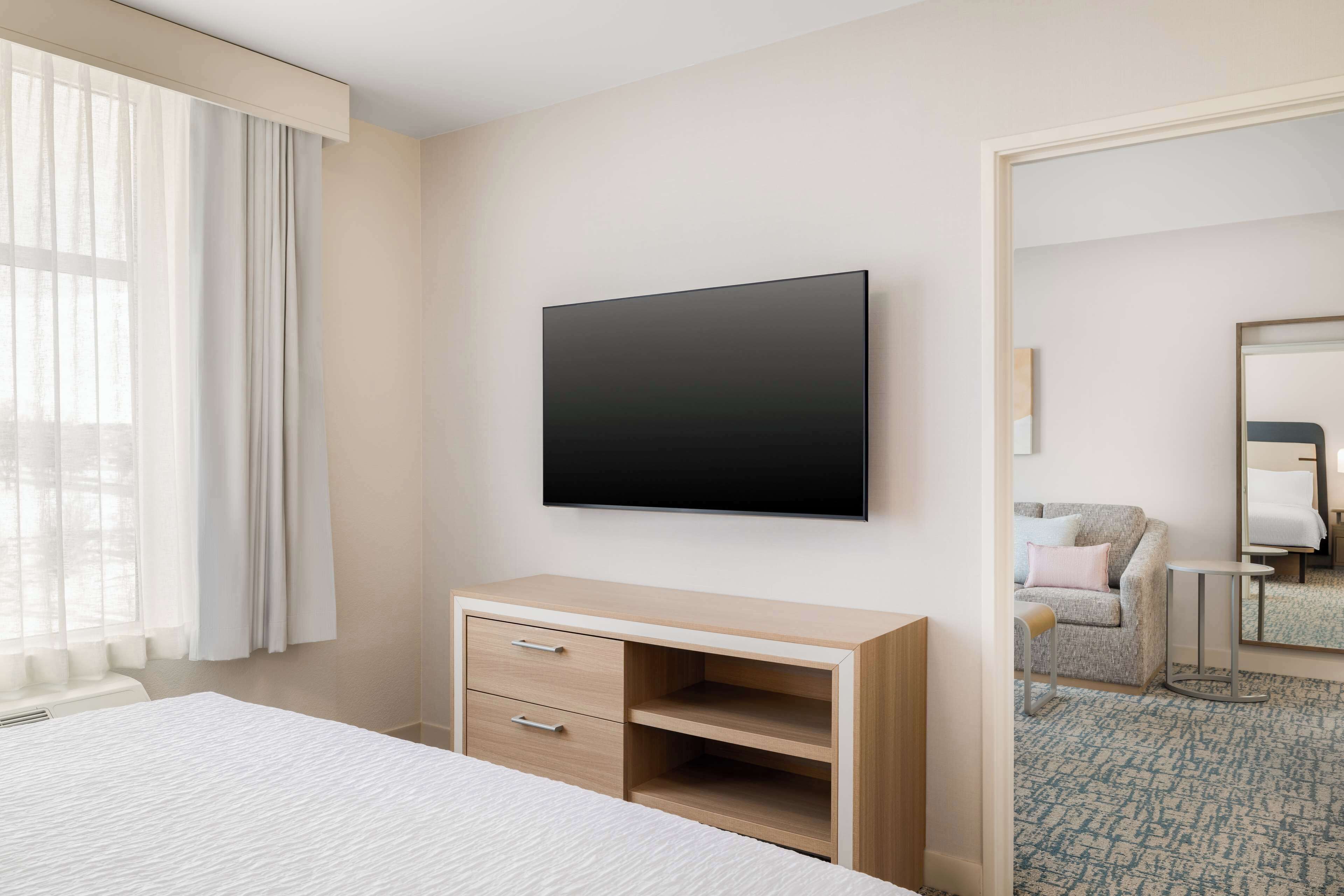 A hotel room with a bed, a desk with drawers, a flat-screen TV mounted on the wall, a window with curtains, and a patterned carpet.