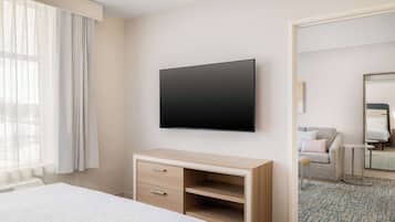 A hotel room with a bed, a desk with drawers, a flat-screen TV mounted on the wall, a window with curtains, and a patterned carpet.