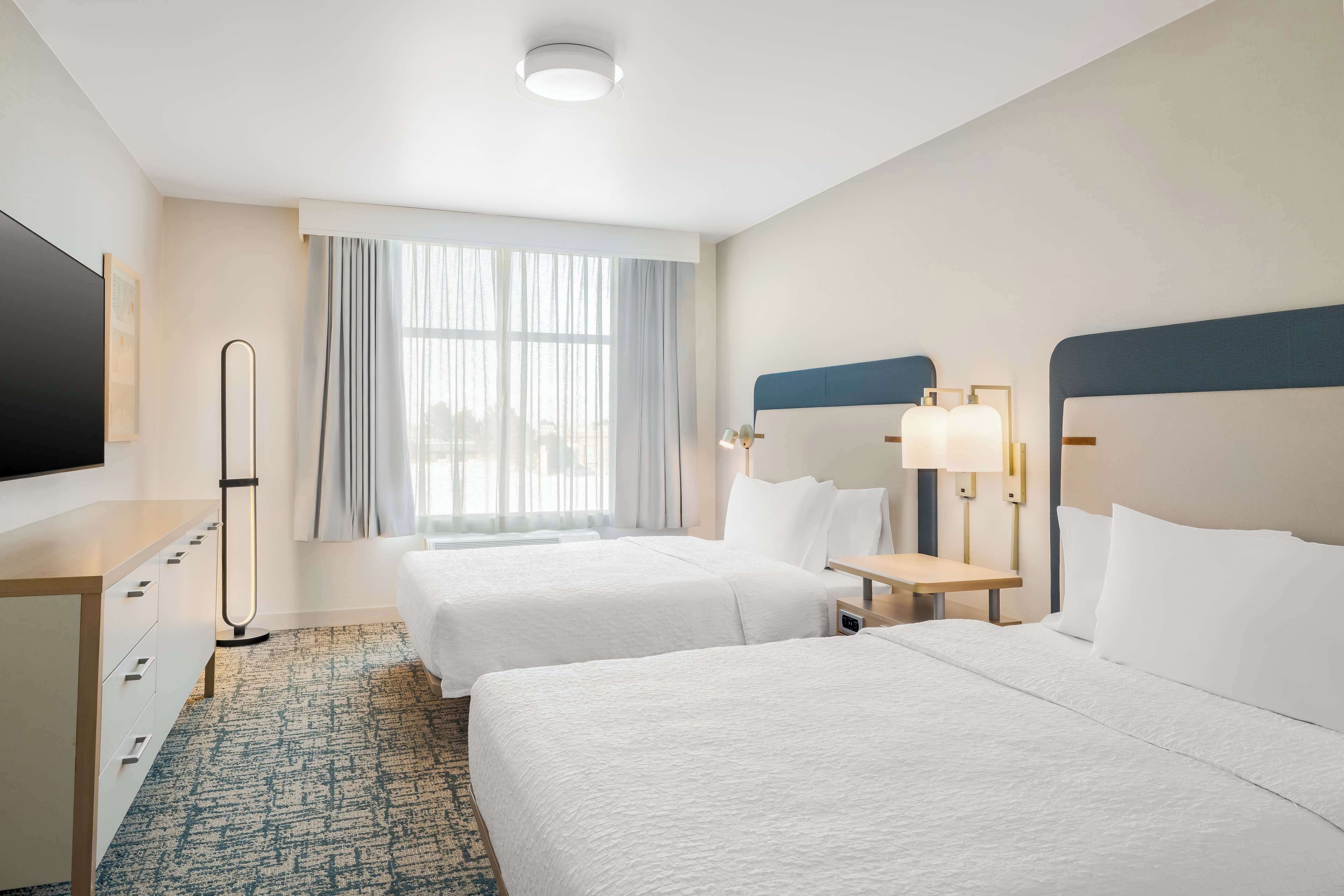 A hotel room with two beds, a desk, a flat-screen TV, and a large window with curtains.