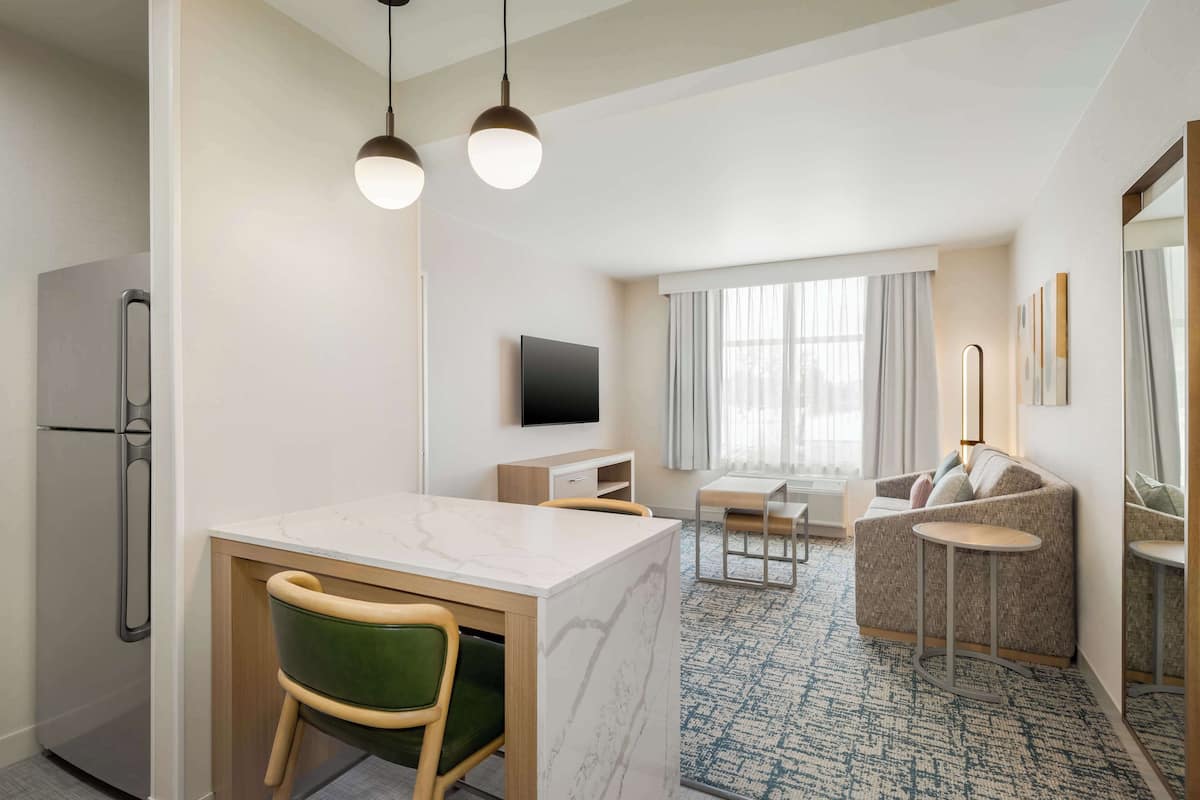 A modern hotel room with a kitchen area, a dining table with chairs, a sofa, and a small round table.