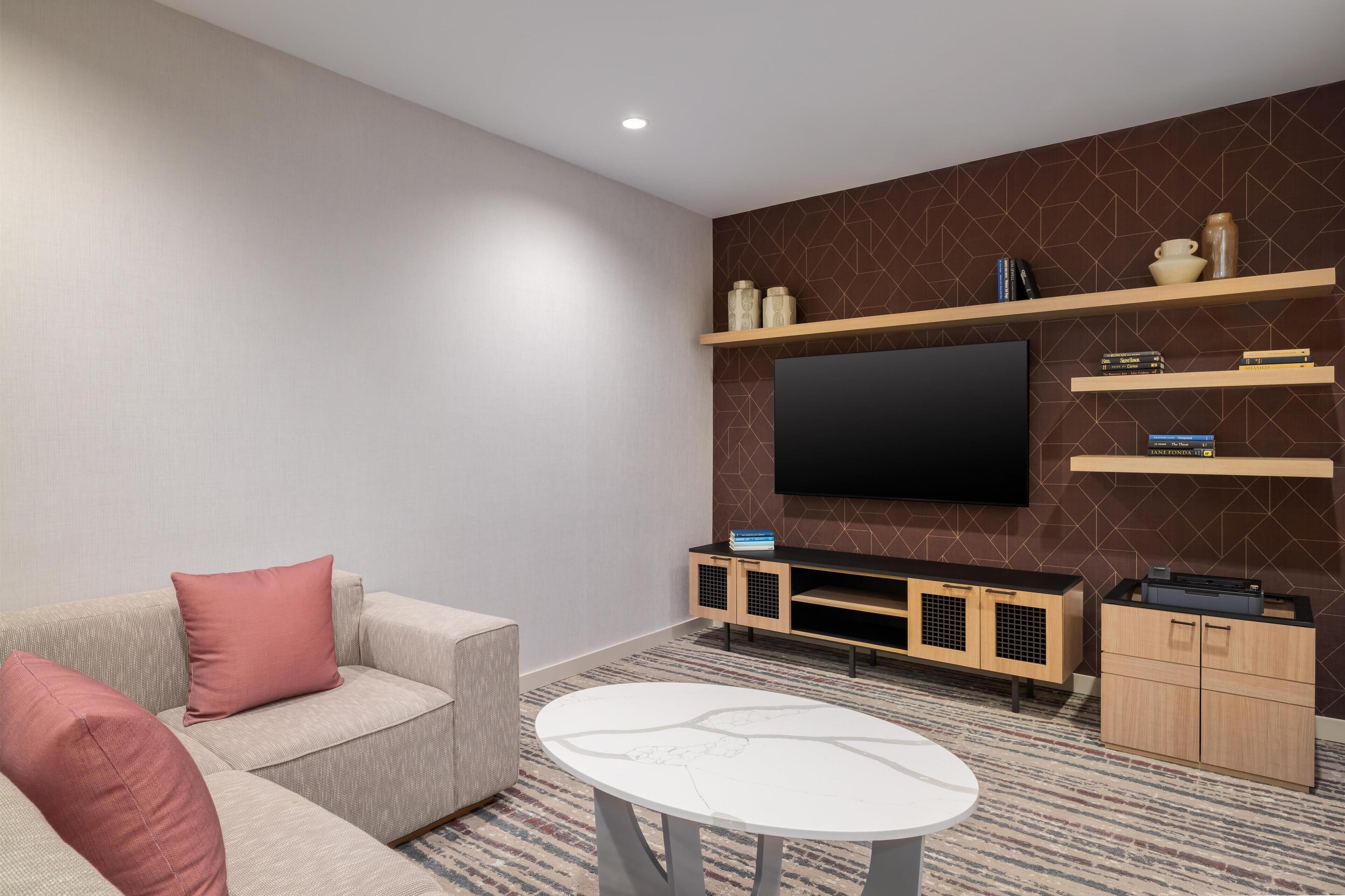 A modern living room with a flat-screen TV mounted on the wall, a beige sofa with pink cushions, a white coffee table, and wooden shelving with decorative items and books.
