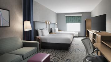 A hotel room with two beds, a desk, a chair, a sofa, a TV, and a lamp.