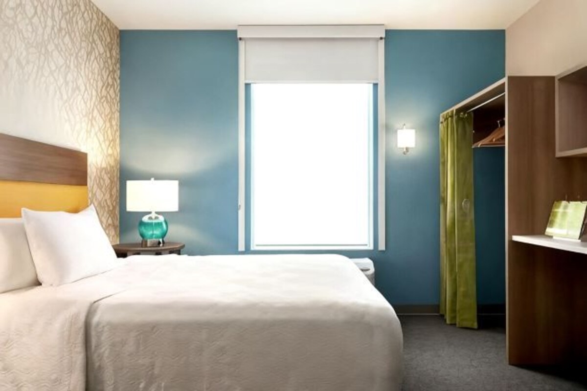 Habitación de hotel con cama, mesita de noche, lámpara, ventana con persiana, armario y estante con objetos.