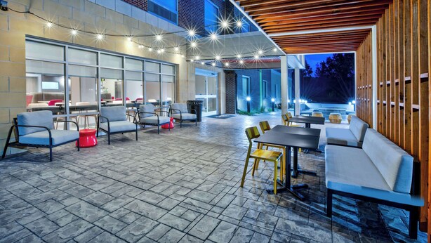 Un patio exterior con asientos y mesas, luces colgantes y vistas al interior del edificio.