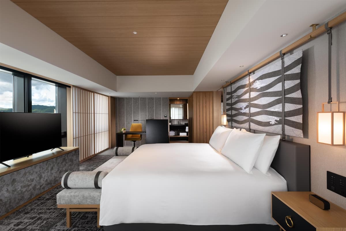 A modern hotel room with a large bed, a flat-screen TV, a desk, and a view of the mountains.