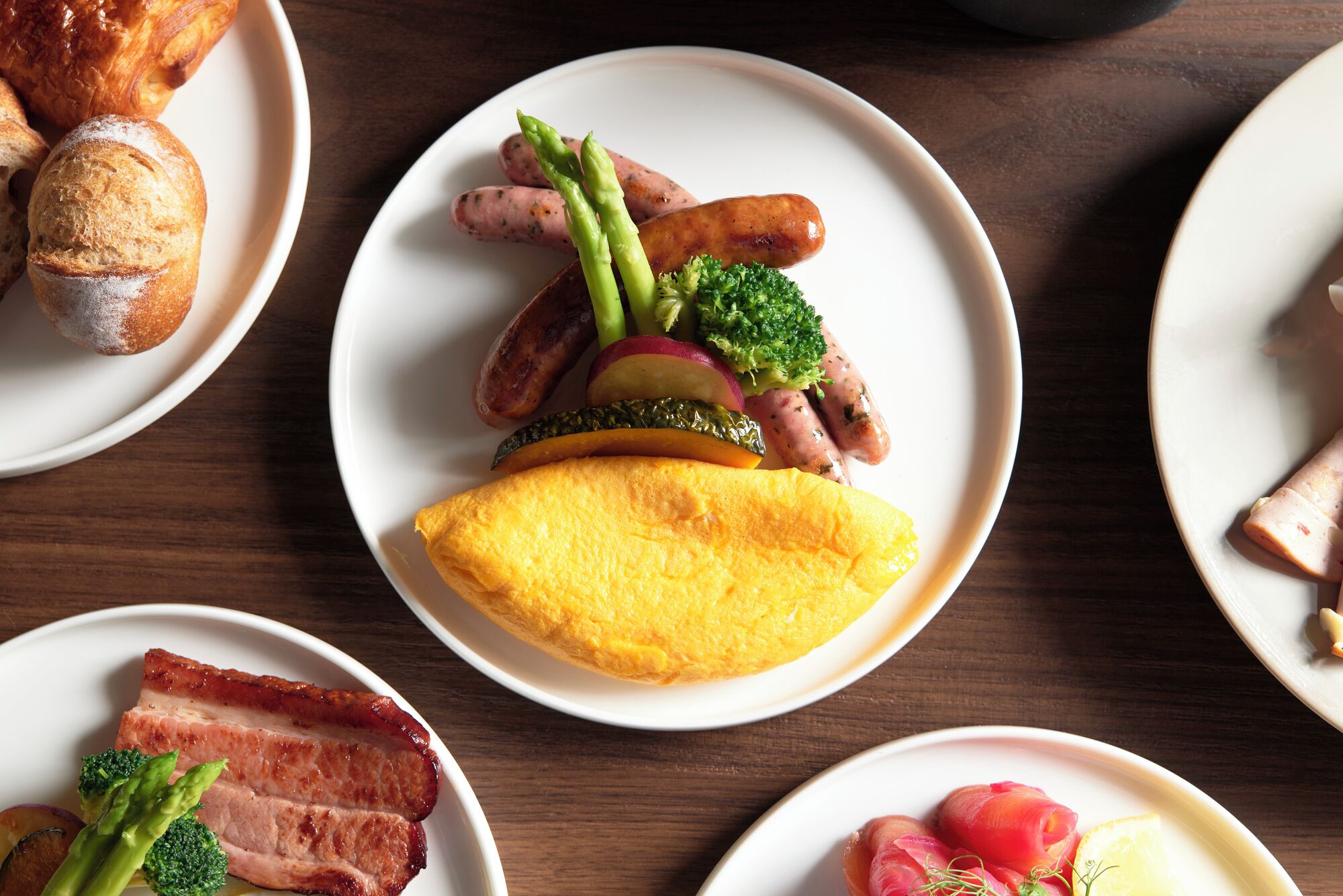 A table with plates of food including sausages, asparagus, broccoli, and a slice of ham.