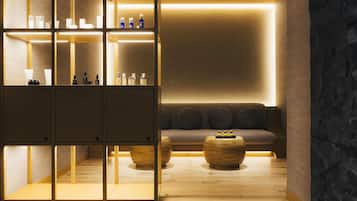 A modern spa room with wooden flooring, a seating area with cushions, and a shelf with various bottles.
