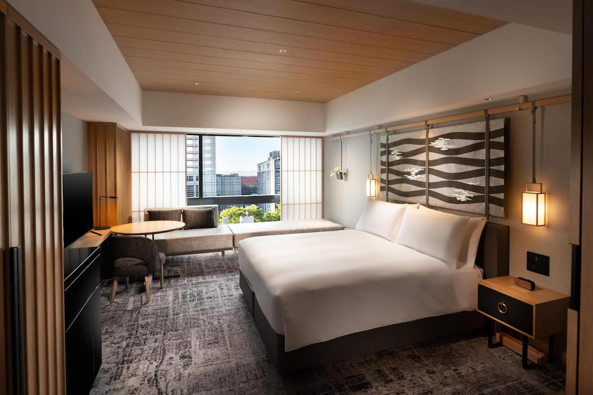 A modern hotel room with a large bed, a desk with a chair, a small table, and a view of the city through large windows.