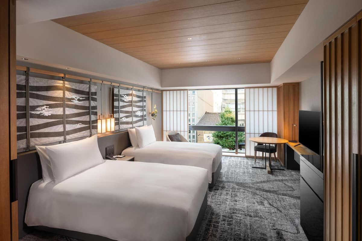 A hotel room with two beds, a TV, a small table, and a view of the city.
