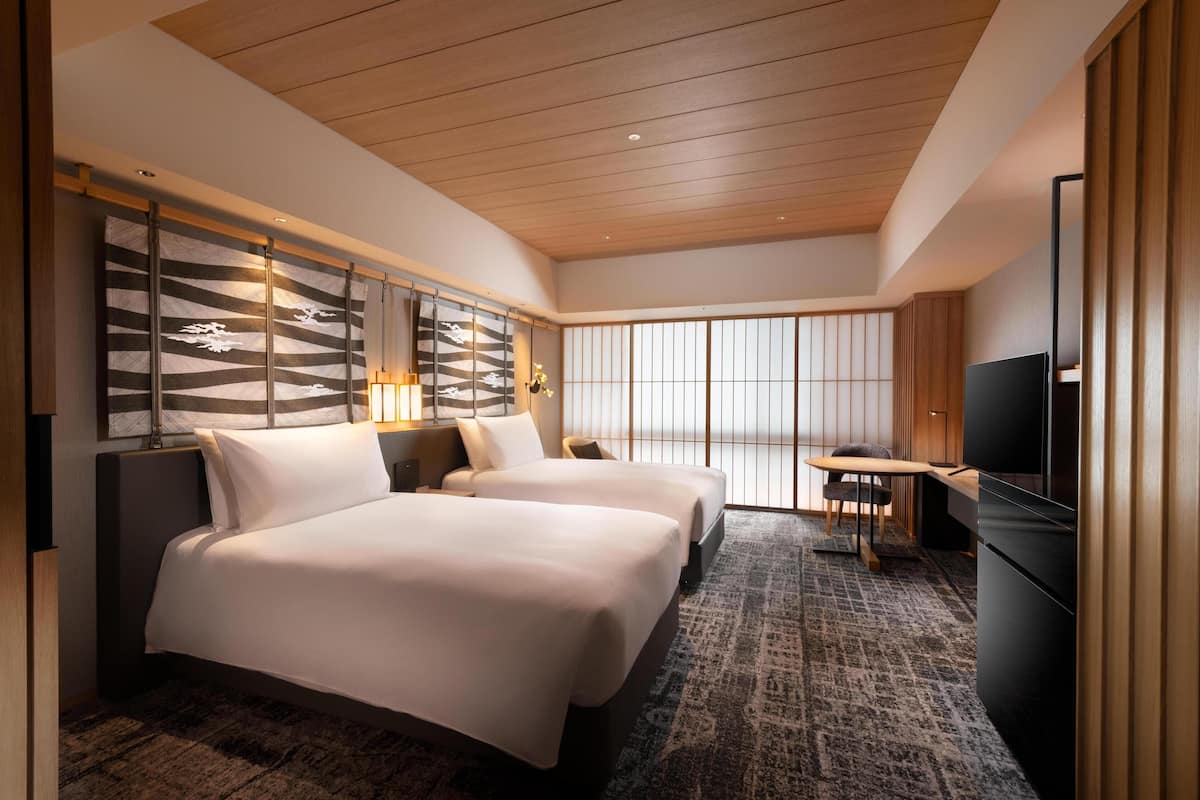 A hotel room with two beds, a TV, a small table, and a view of the outside through large windows.