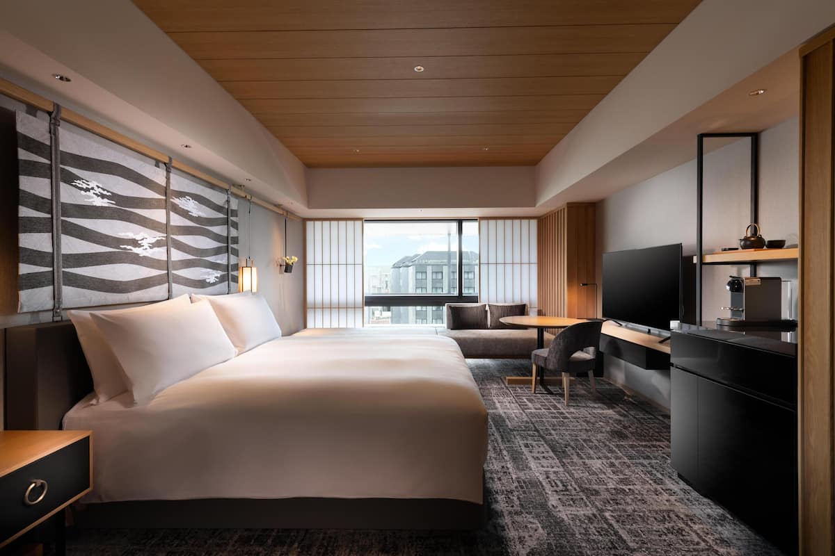 A modern hotel room with a large bed, a desk, a TV, and a view of the city.