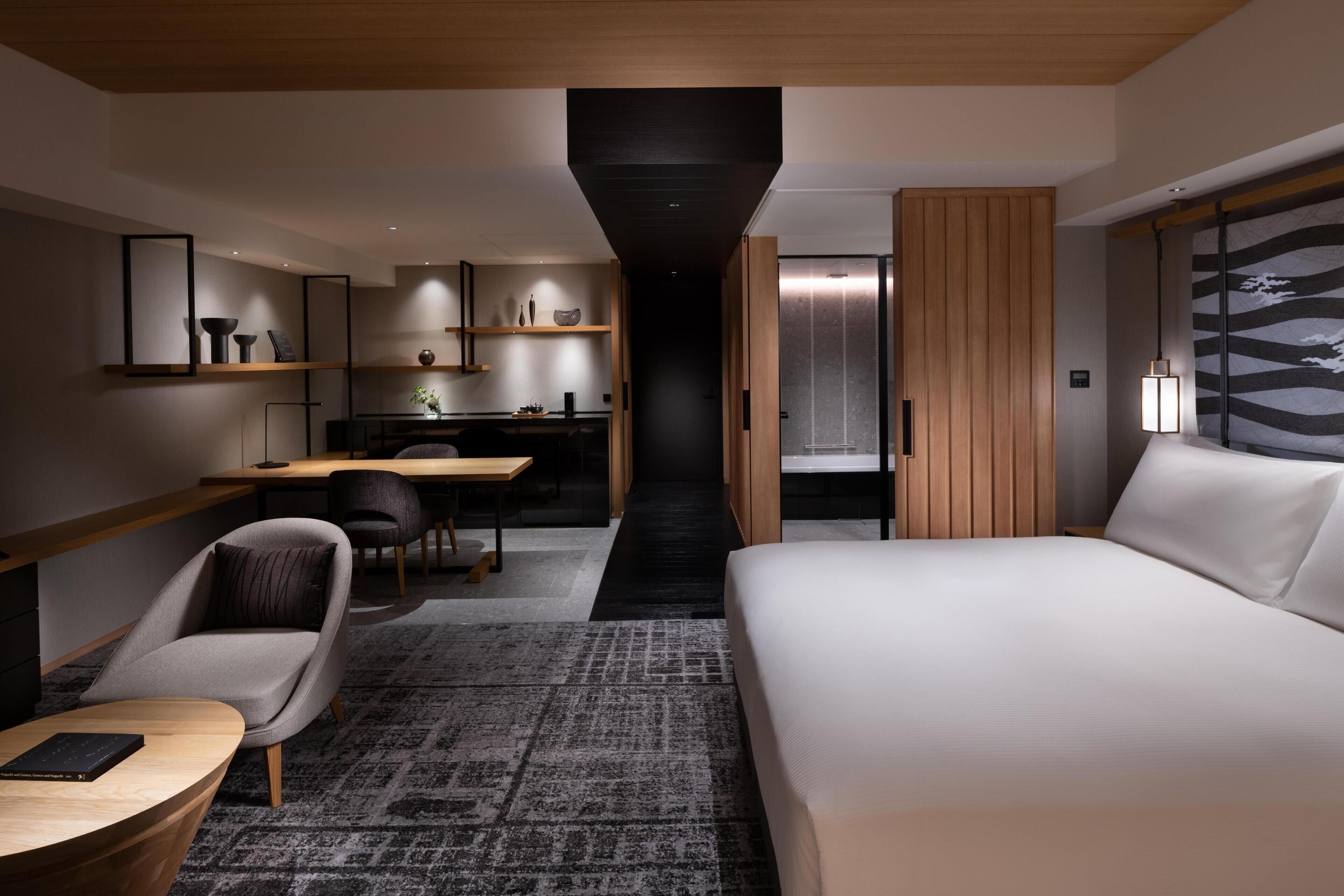A modern hotel room with a large bed, a desk, a chair, and a small table. There is a wardrobe and a mirror on the wall.