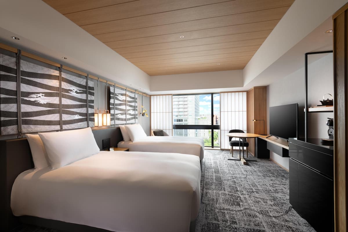 A modern hotel room with two beds, a desk, a TV, and large windows offering a city view.