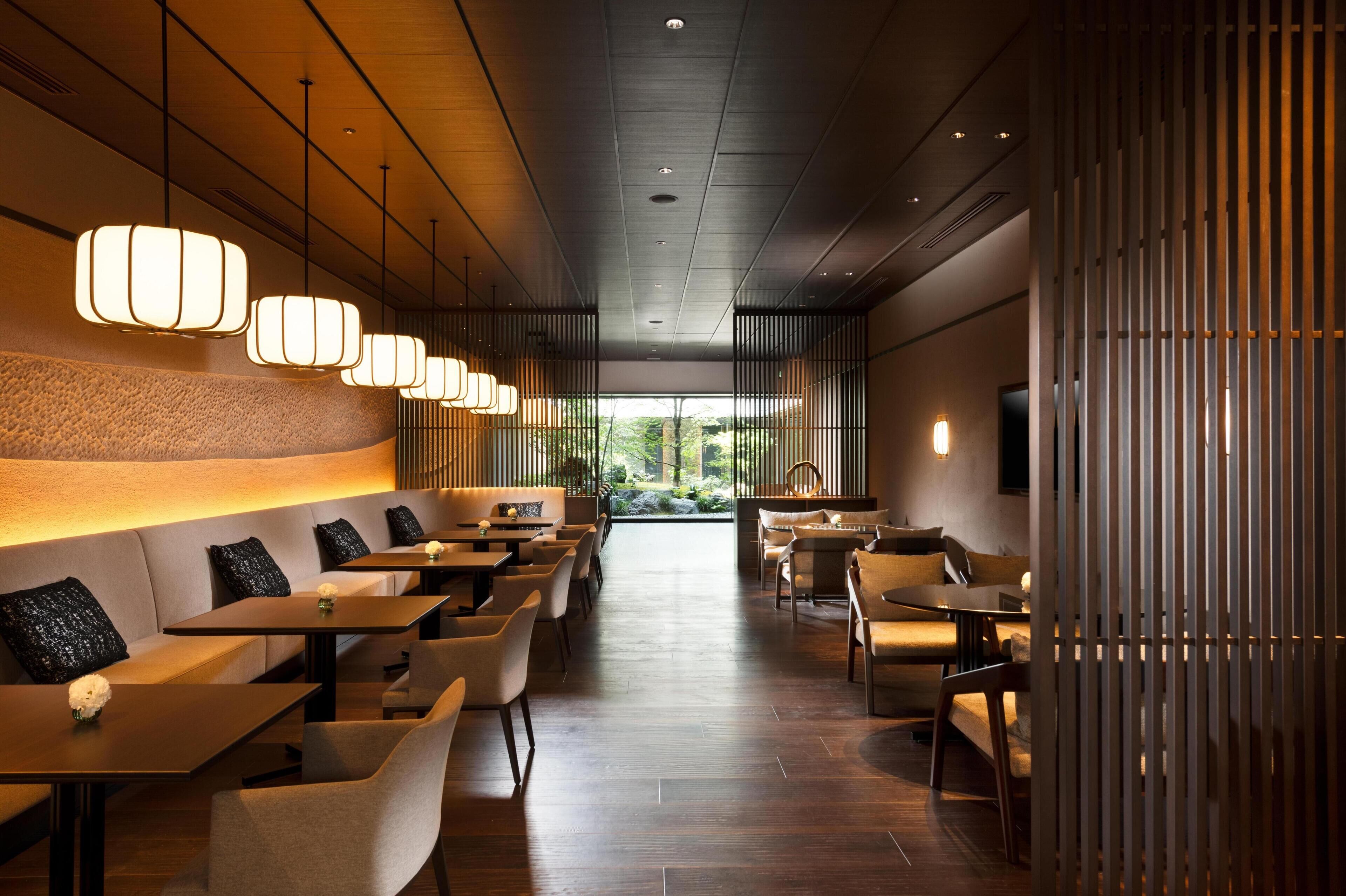 A modern restaurant interior with a long counter, cushioned seating, pendant lighting, and a view of greenery outside.