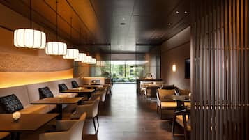 A modern restaurant interior with a long counter, cushioned seating, pendant lighting, and a view of greenery outside.
