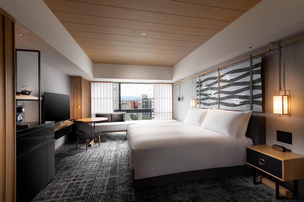 A modern hotel room with a large bed, a desk, a TV, and a view of the city.