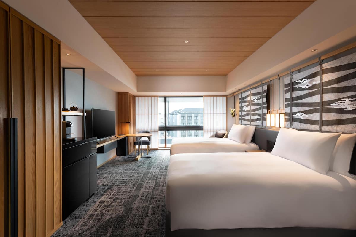 A modern hotel room with a large bed, a desk with a television, a small table, and a view of the city through large windows.