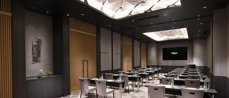 A conference room with a geometric ceiling design, multiple tables with chairs, and a large screen displaying the Hilton logo.