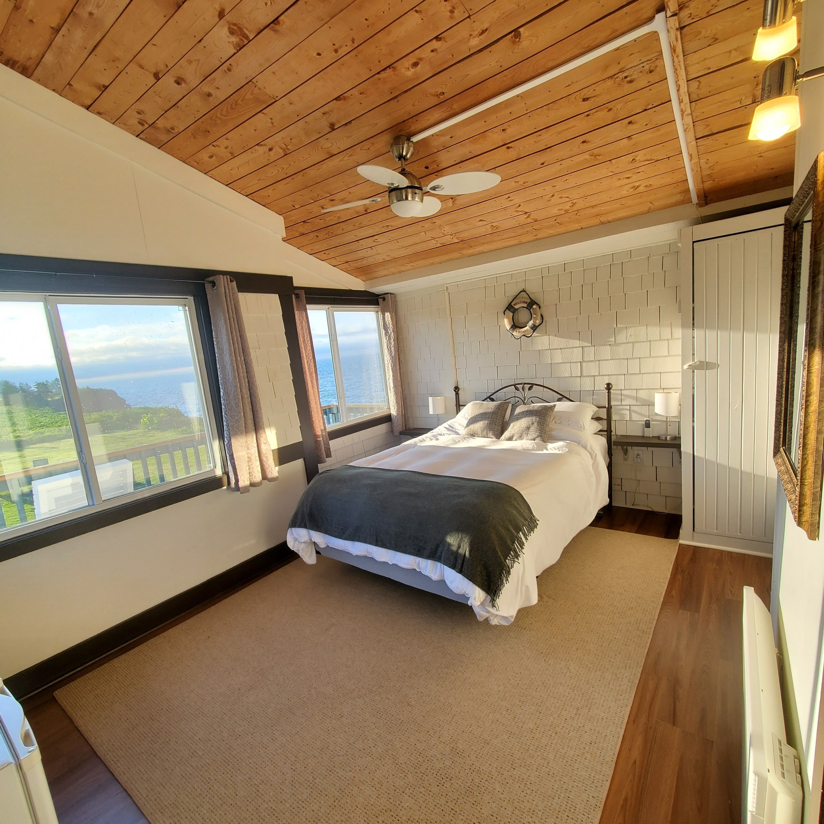A bedroom with a bed, a ceiling fan, a view of the sea, and a white tiled wall.