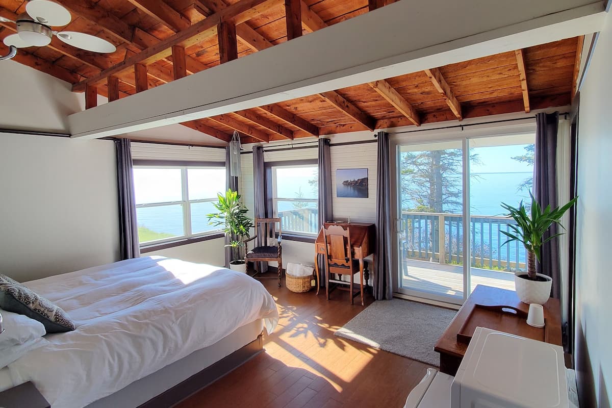 A bedroom with a large bed, a desk, a chair, and a view of the sea through large windows.