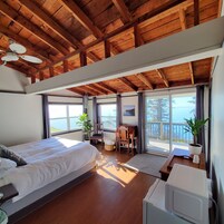 A bedroom with a large bed, a desk, a chair, and a view of the sea through large windows.