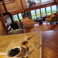 A cozy living room with a wooden floor, a glass table with a wine glass and a bowl of mussels, and a view of a garden through large windows.