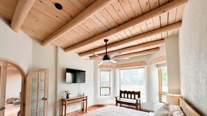 Un dormitorio con una cama grande, vigas de madera en el techo, un ventilador de techo, un televisor montado en la pared y una ventana con vistas al exterior.