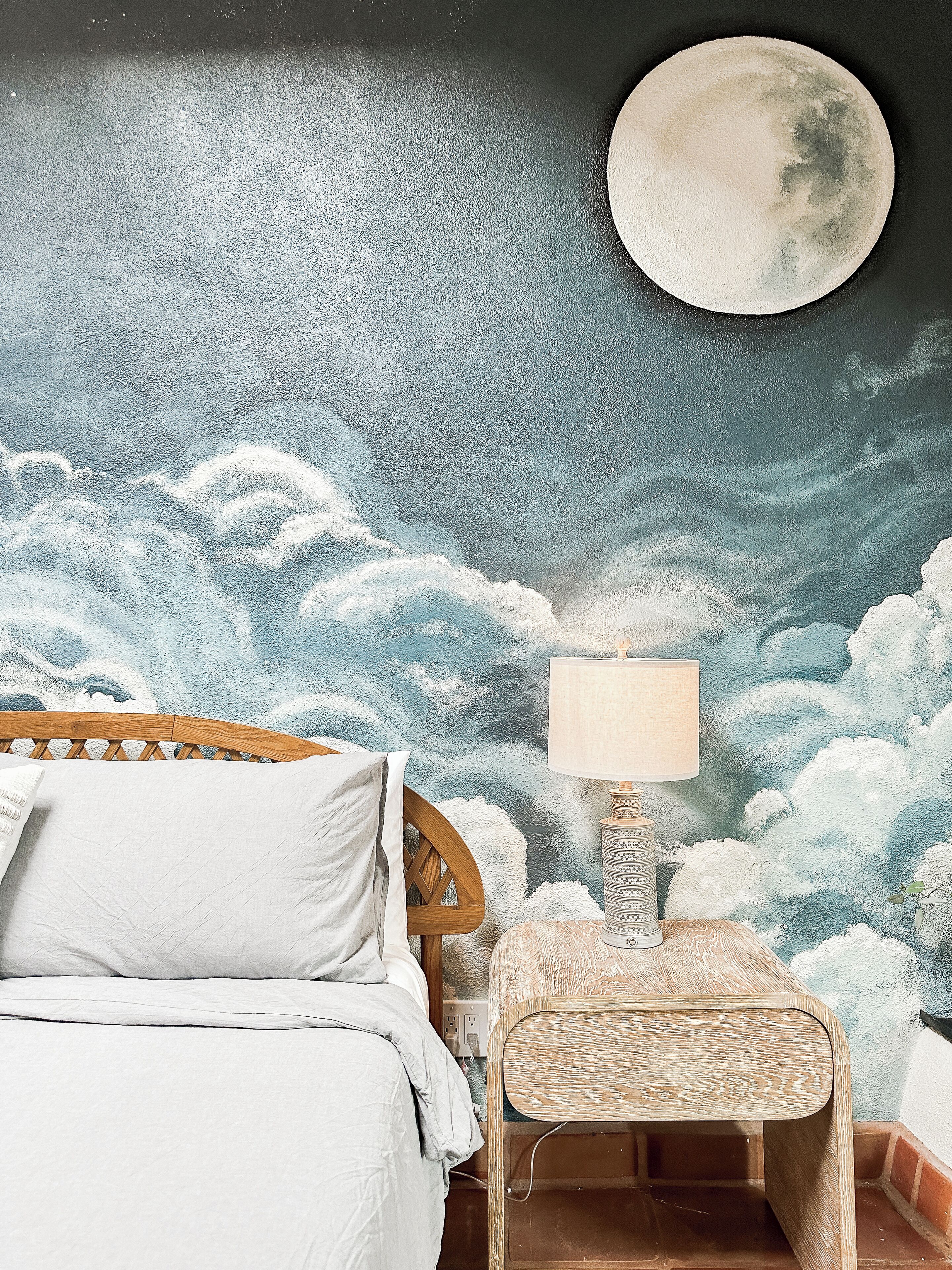 Un dormitorio con cama, mesita de noche, lámpara y un mural en la pared con nubes y luna.