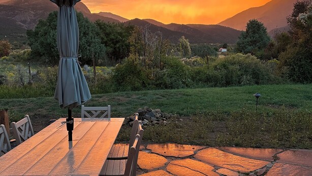 Mesa y sillas de madera en un patio de piedra con sombrilla, frente a un atardecer sobre un paisaje montañoso.