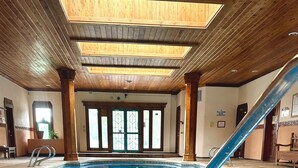 Una piscina interior con agua azul cristalina, vigas de madera en el techo y vistas al exterior a través de grandes ventanales.