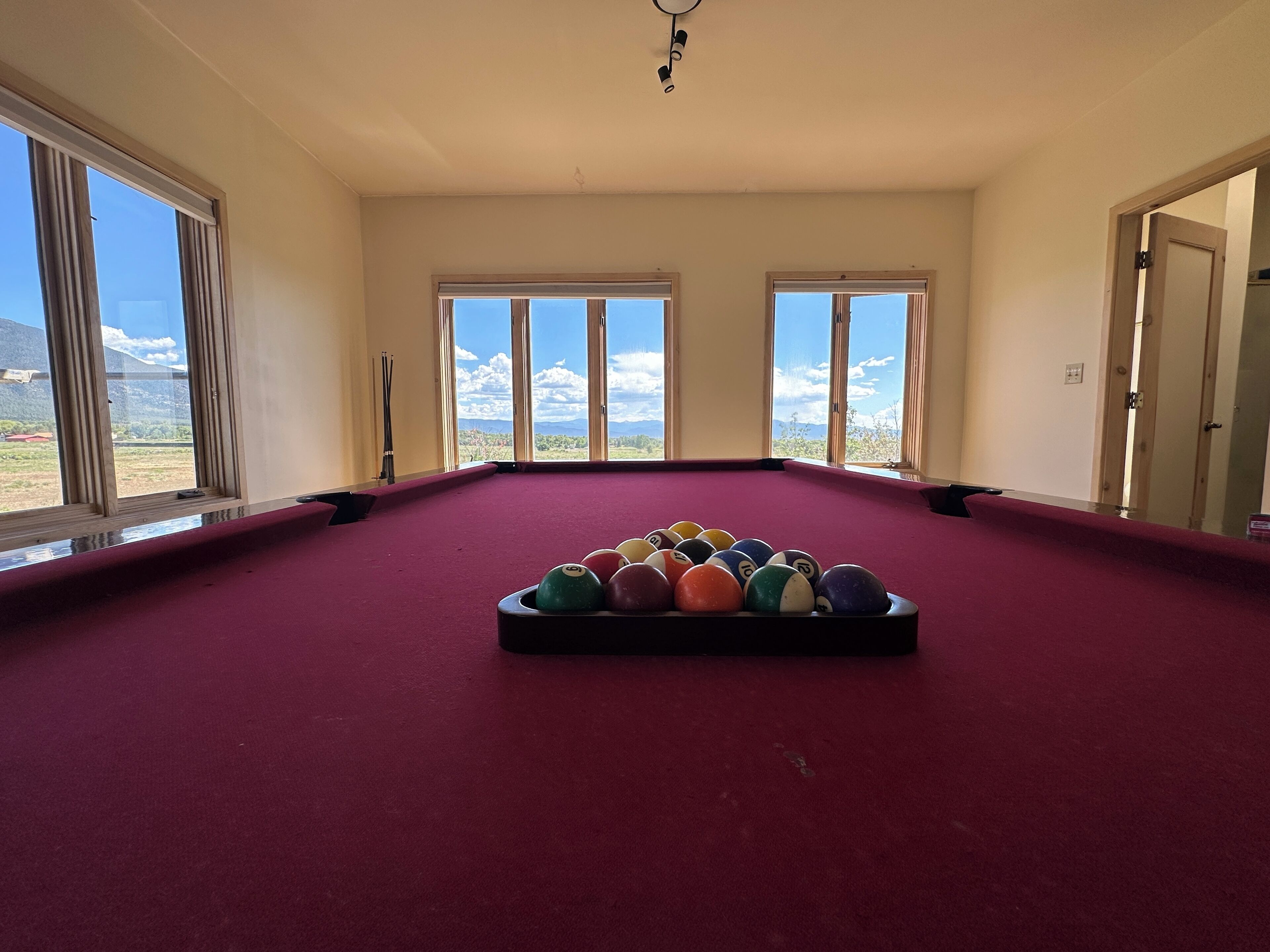 A pool table with balls in the center, large windows with a view of mountains, and a door on the right.