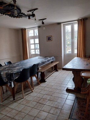 Zona pranzo con un tavolo lungo, panche e sedie. Sono presenti finestre con tende, una ventola a soffitto e un quadro appeso al muro.