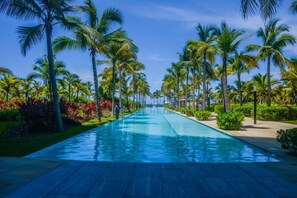 A long, narrow swimming pool with palm trees on both sides, surrounded by well-maintained greenery. - Península 4-C 03 Recamaras vista al mar (Ixtapa)