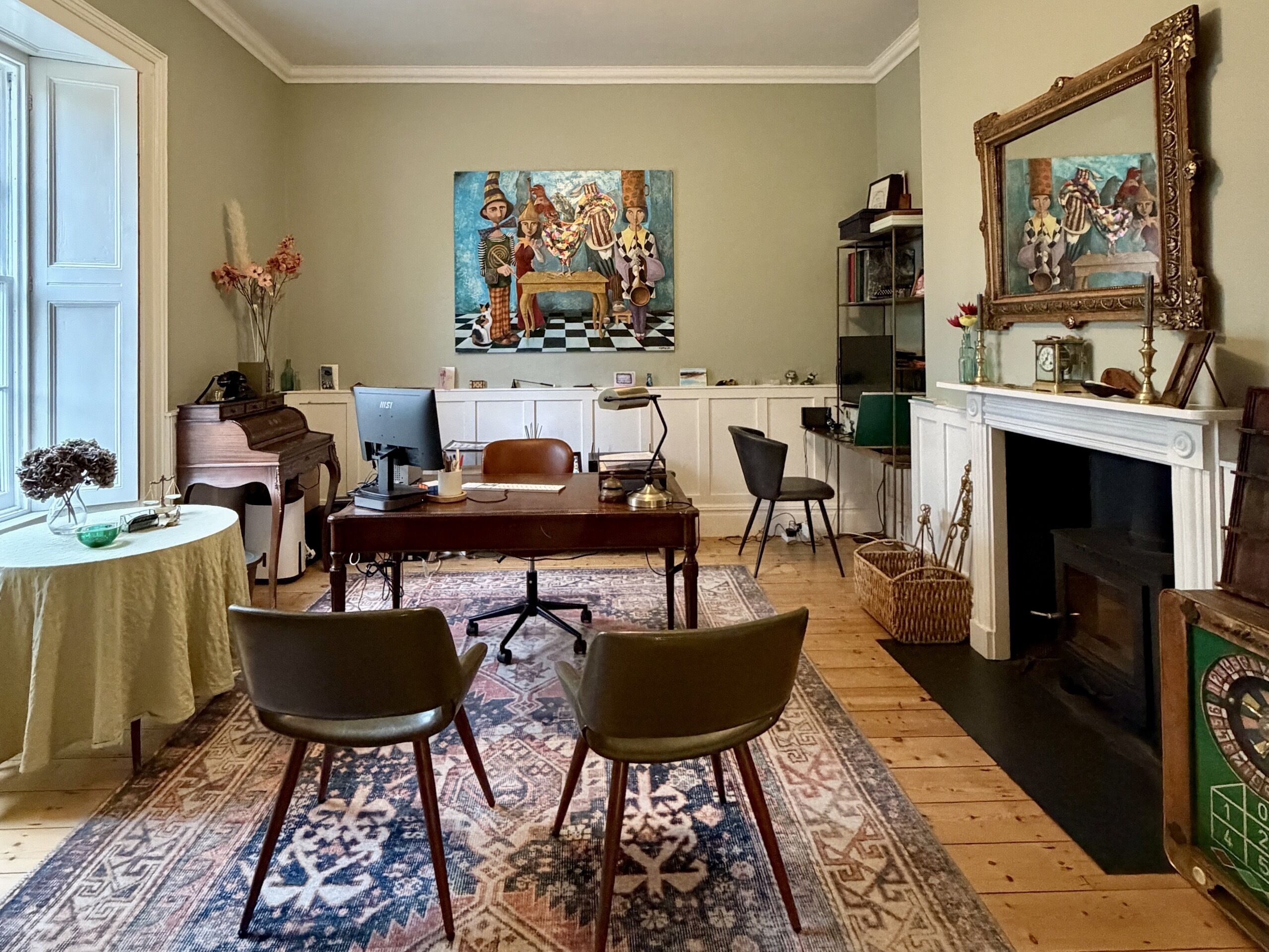 A room with a dining table, two chairs, a piano, a fireplace, and a painting on the wall.