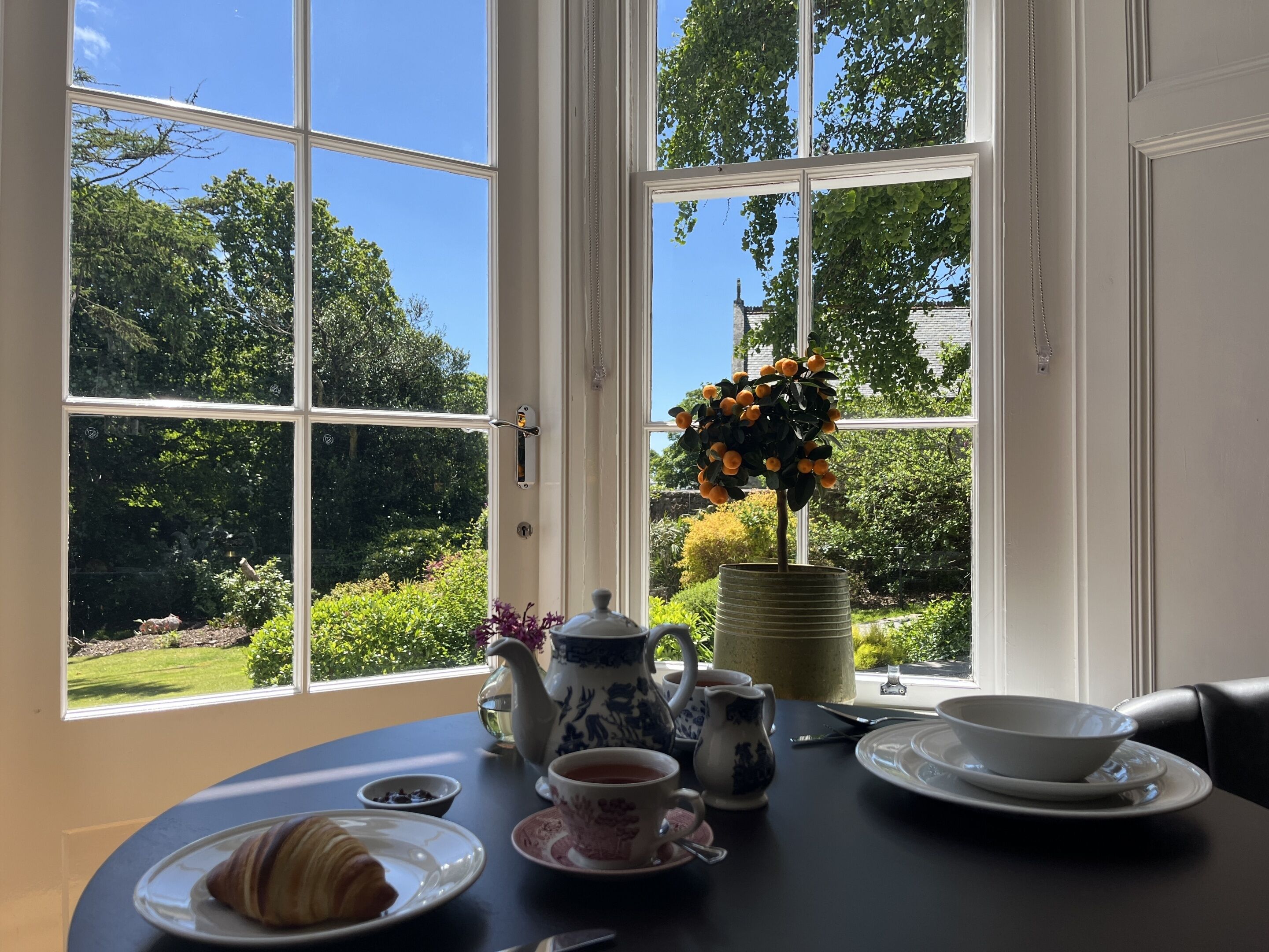 Una mesa con un croissant, tetera, tazas y un cuenco, lista para el té, con vista a un jardín y un árbol con frutos naranjas.