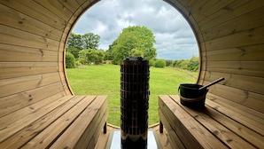 Una estructura circular de madera con una chimenea central, bancos y vistas a una zona cubierta de césped y árboles.