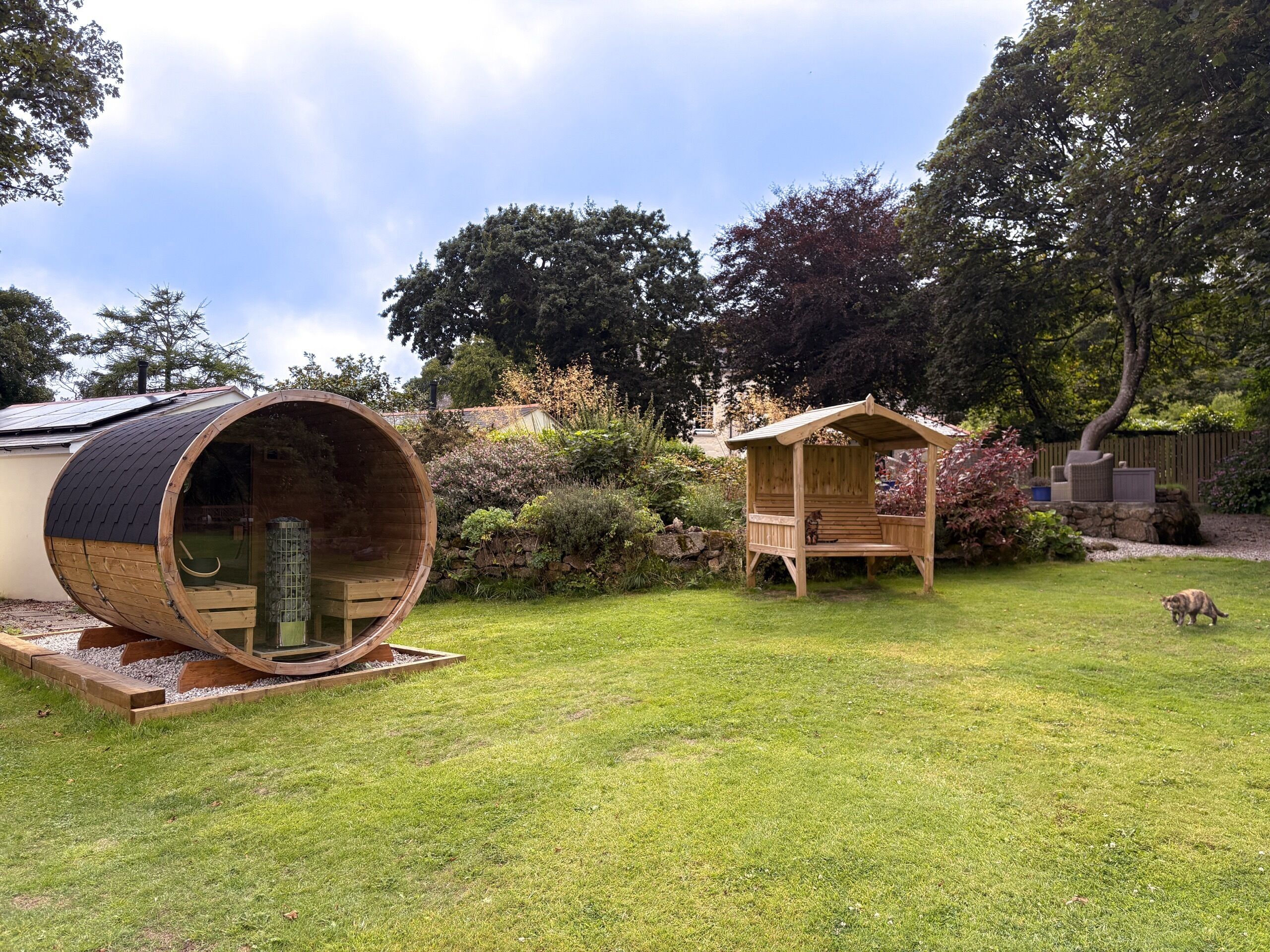 Una estructura de sauna de madera con techo inclinado, un banco de madera y una pequeña cabaña de madera con techo a dos aguas en un jardín.