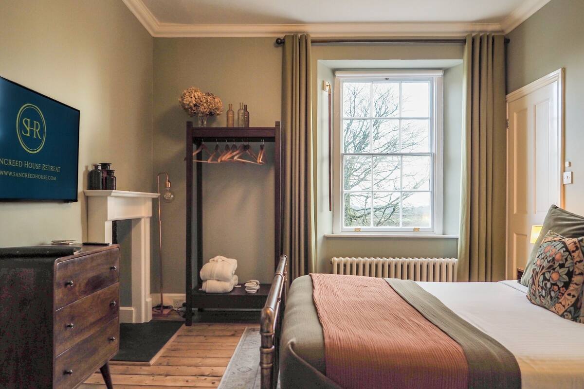 Ein Schlafzimmer mit Bett, Schminktisch, Kleiderhaken, einem Fenster mit Vorhängen und einem an der Wand befestigten Schild.