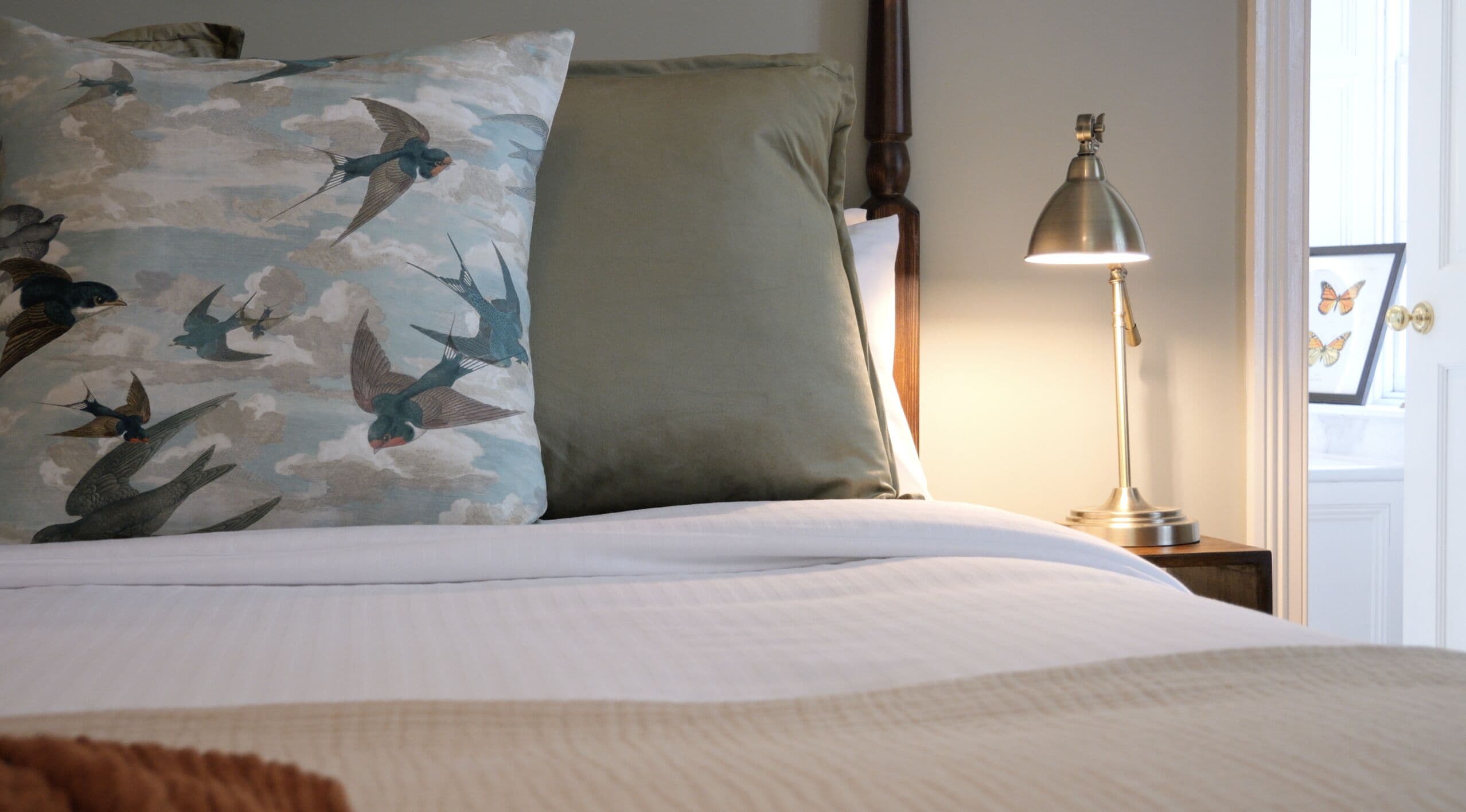 Una cama bien hecha con ropa blanca, un almohadón con estampado de pájaro, un almohadón verde y una lámpara de noche sobre una mesita de madera.