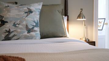 Una cama bien hecha con ropa blanca, un almohadón con estampado de pájaro, un almohadón verde y una lámpara de noche sobre una mesita de madera.