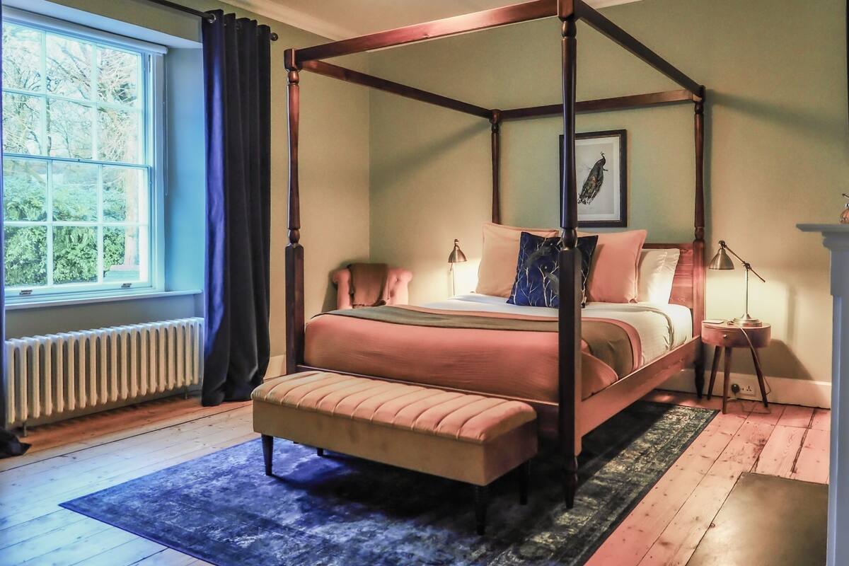 Ein Himmelbett mit Sitzbank, ein Heizkörper, ein Fenster mit Vorhängen und ein gerahmtes Bild an der Wand.