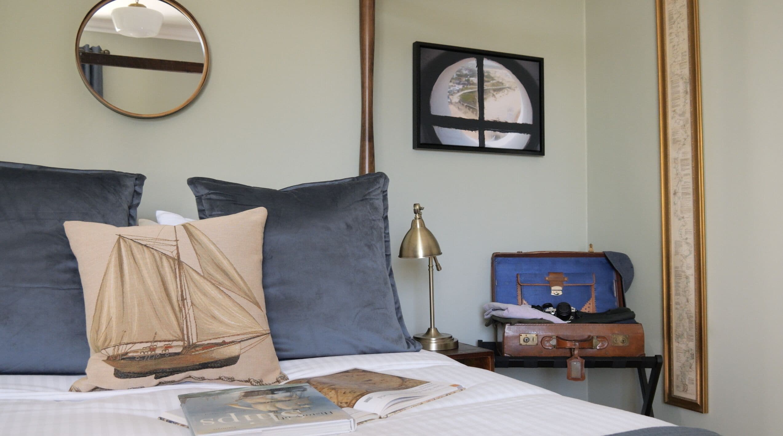 A neatly made bed with pillows, a book, a lamp, and a suitcase on a nightstand.