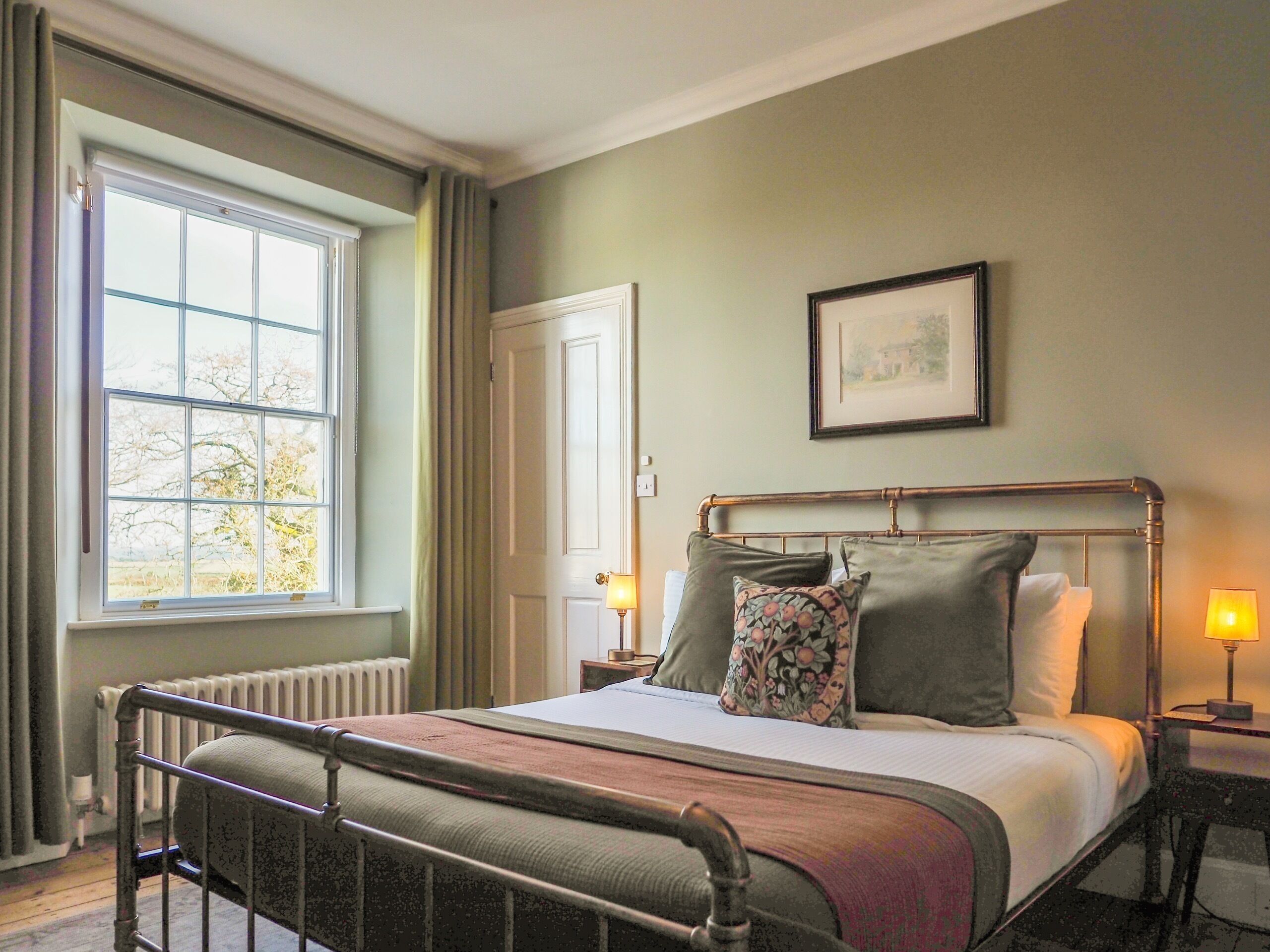 A bedroom with a metal bed frame, a large window with curtains, a bedside table with a lamp, and a framed picture on the wall.