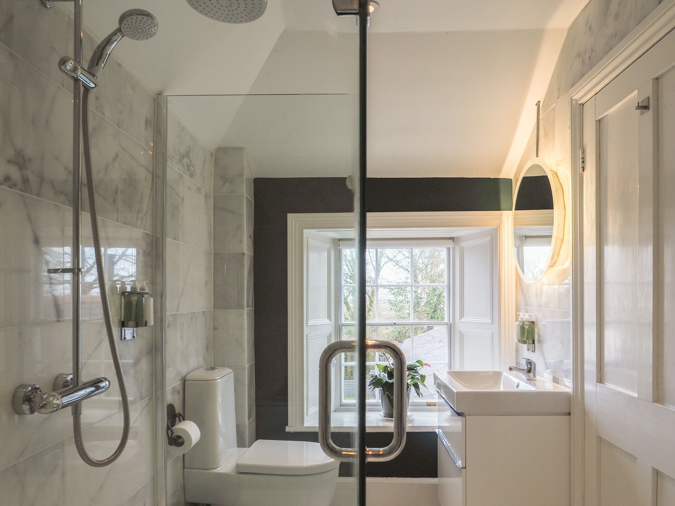 A bathroom with a shower, toilet, sink, and a window with a view of greenery.