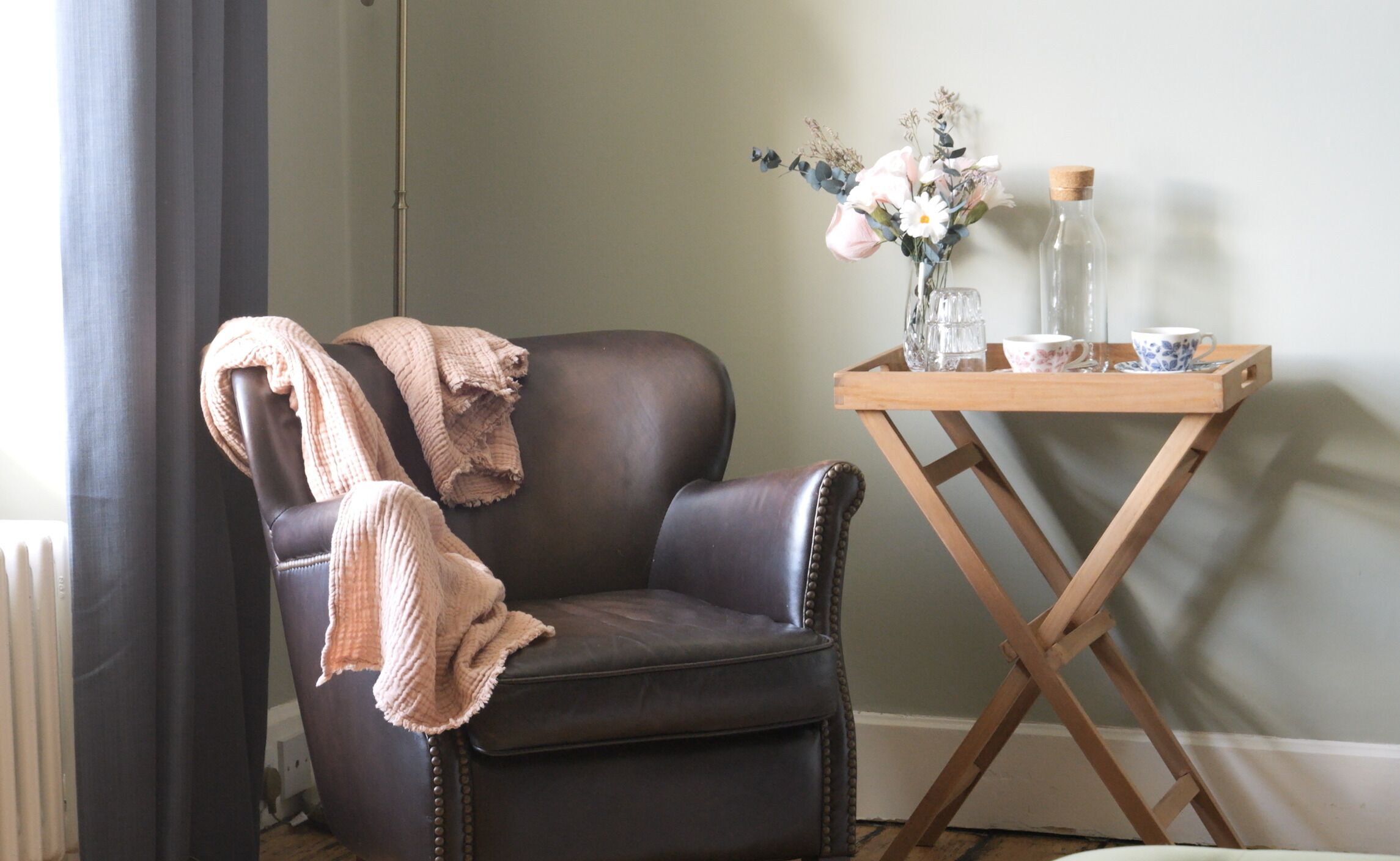 A dark brown leather armchair with a pink knitted throw, a wooden side table with a vase of flowers, a glass bottle, and a cup.