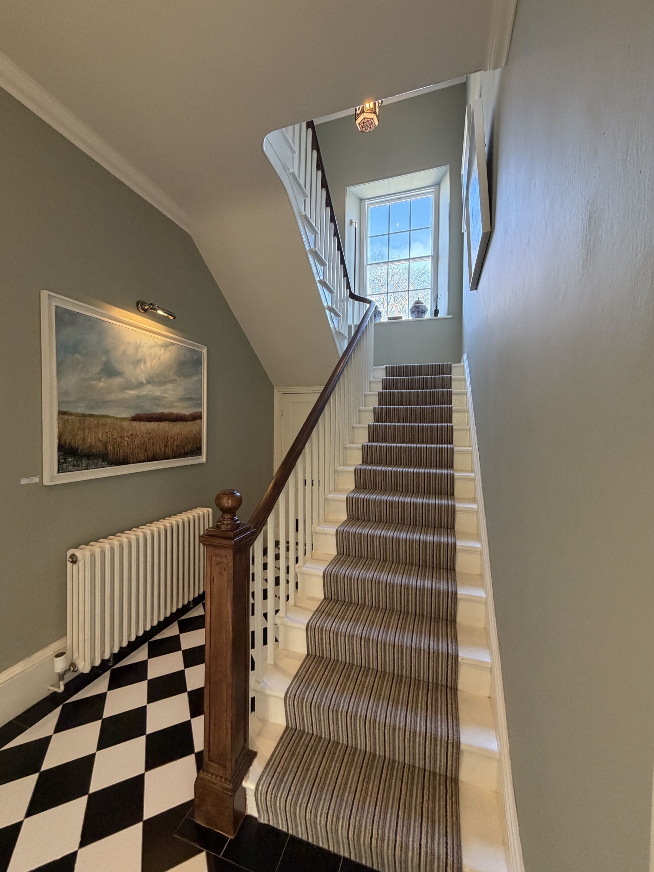 Un escalier avec un sol à carreaux, un radiateur et un tableau représentant un paysage, encadré.