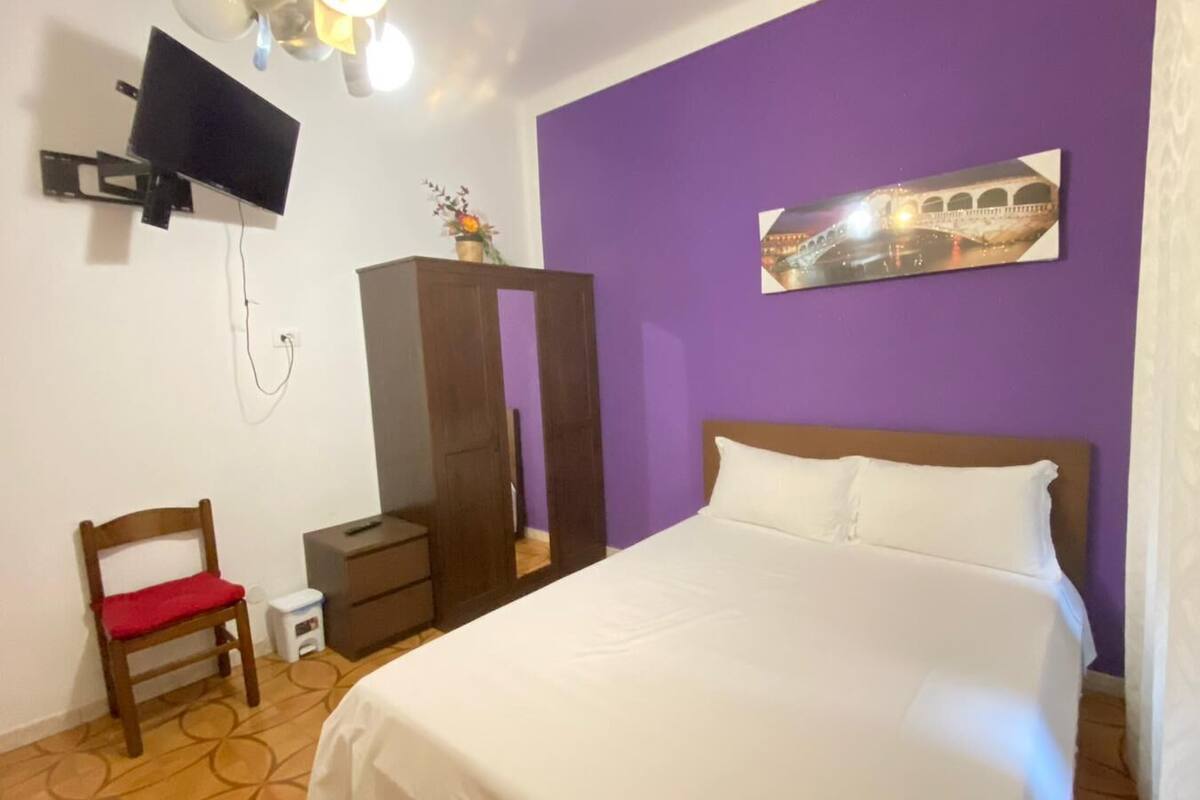 A bedroom with a bed, a TV mounted on the wall, a wardrobe, and a small table with a chair.