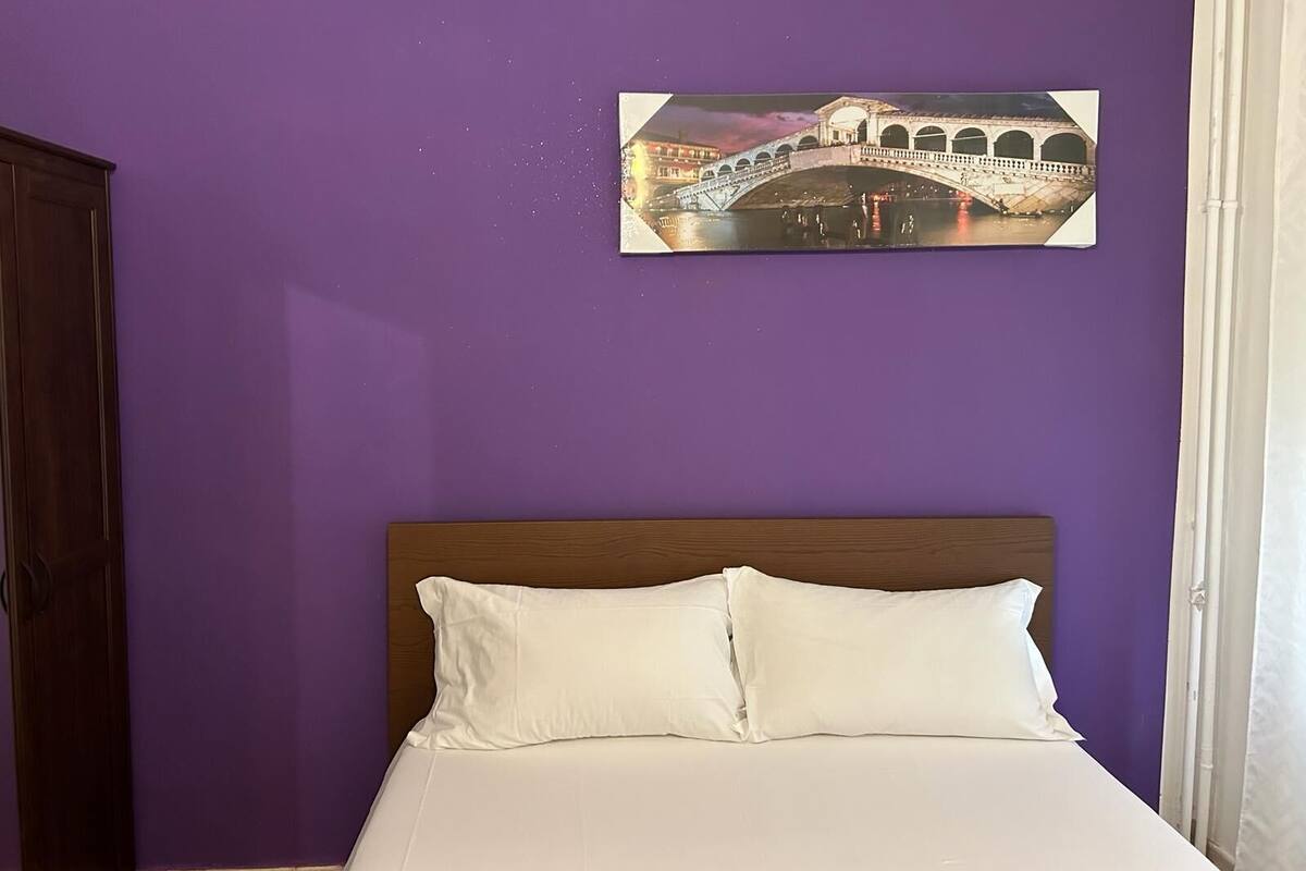 A bedroom with a bed, a wooden headboard, a purple wall, and a framed picture of a bridge.